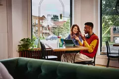 Female smiling towards a male with headphones, sitting next to each other at a table with standing tablet, in a cafe