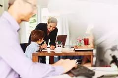 Smiling female helping her kid draw at the kitchen table. male working on a computer in the foreground.
