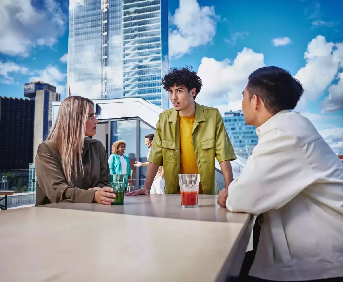 Male in yellow shirt, female and male, standing outside on a balcony at a table, talking