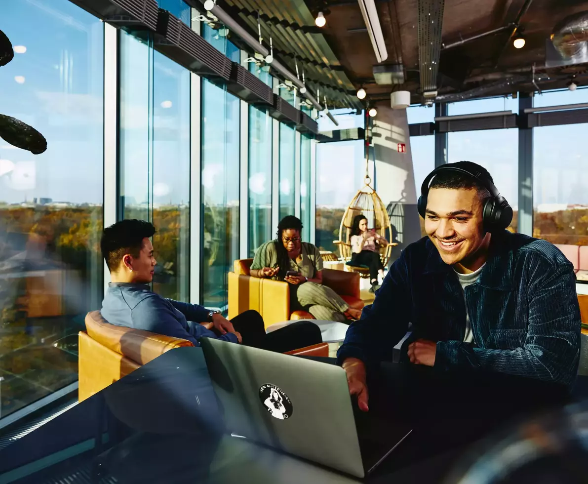 Smiling male with headphones and laptop sitting in a lounge environment.
